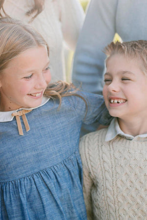 Two children are smiling at each other; one wears a chambray dress with a Peter Pan collar and bow detail, while the other is dressed in a cream cable-knit sweater.