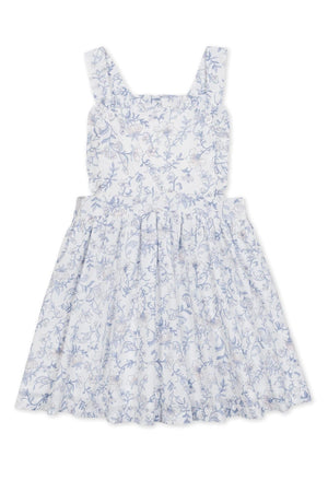 A charming white apron dress features a delicate blue floral pattern, ruffle straps, and a flared skirt. Made from organic cotton, this dress is perfect for girls and toddlers.