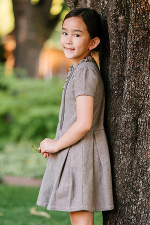 A young girl stands by a tree, wearing a beige and grey houndstooth dress with short sleeves and a ruffle.
