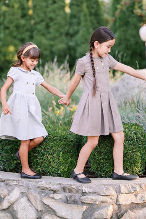 Two young girls in stylish dresses play on a stone pathway, surrounded by greenery, wearing black shoes and smiling joyfully.