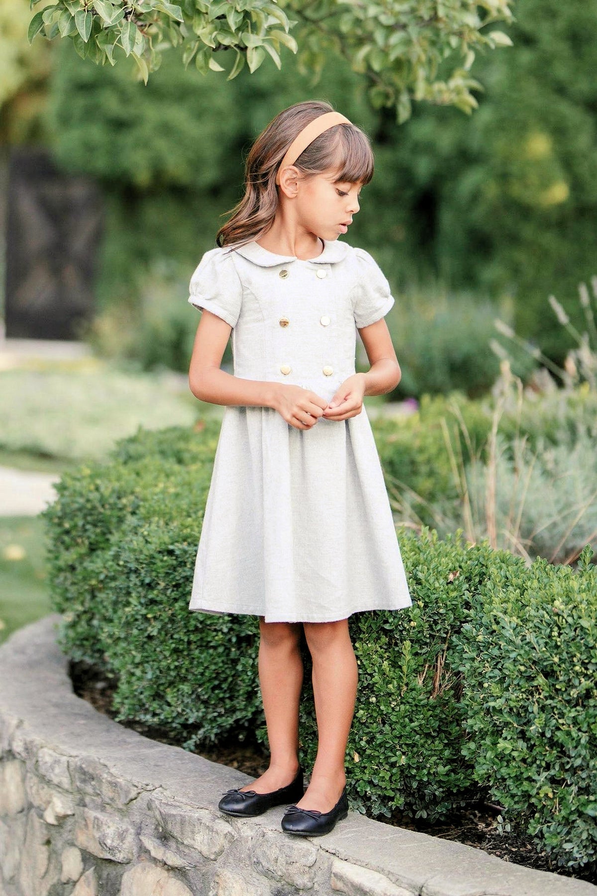 A young girl wears a gray heather flannel dress with puff sleeves and gold buttons, standing beside a lush green.