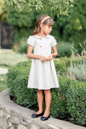 A young girl wears a gray heather flannel dress with puff sleeves and gold buttons, standing beside a lush green.