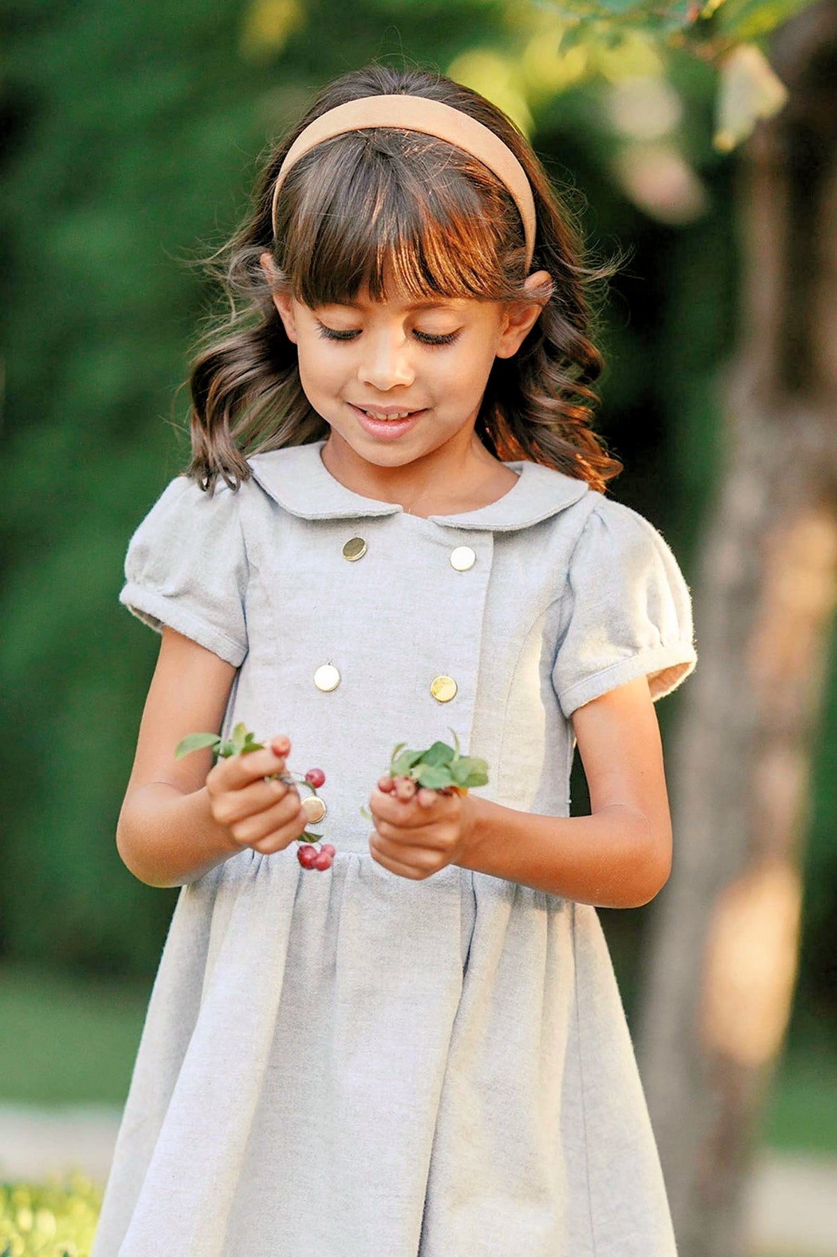 A young girl wearing a gray heather dress with gold buttons holds berries and leaves, smiling in a green outdoor.