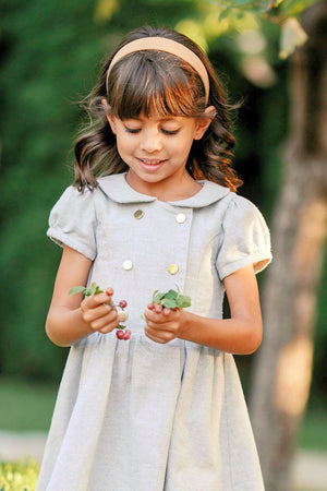 A young girl wearing a gray heather dress with gold buttons holds berries and leaves, smiling in a green outdoor.