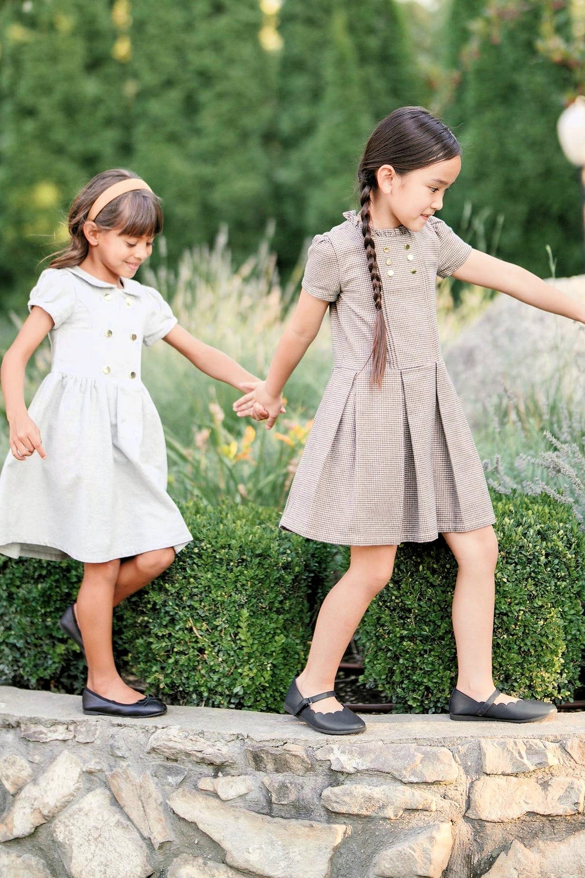 Two young girls hold hands while walking on a stone path, wearing gray and brown dresses and black shoes in.