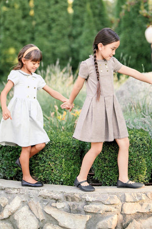 Two young girls hold hands while walking on a stone path, wearing gray and brown dresses and black shoes in.