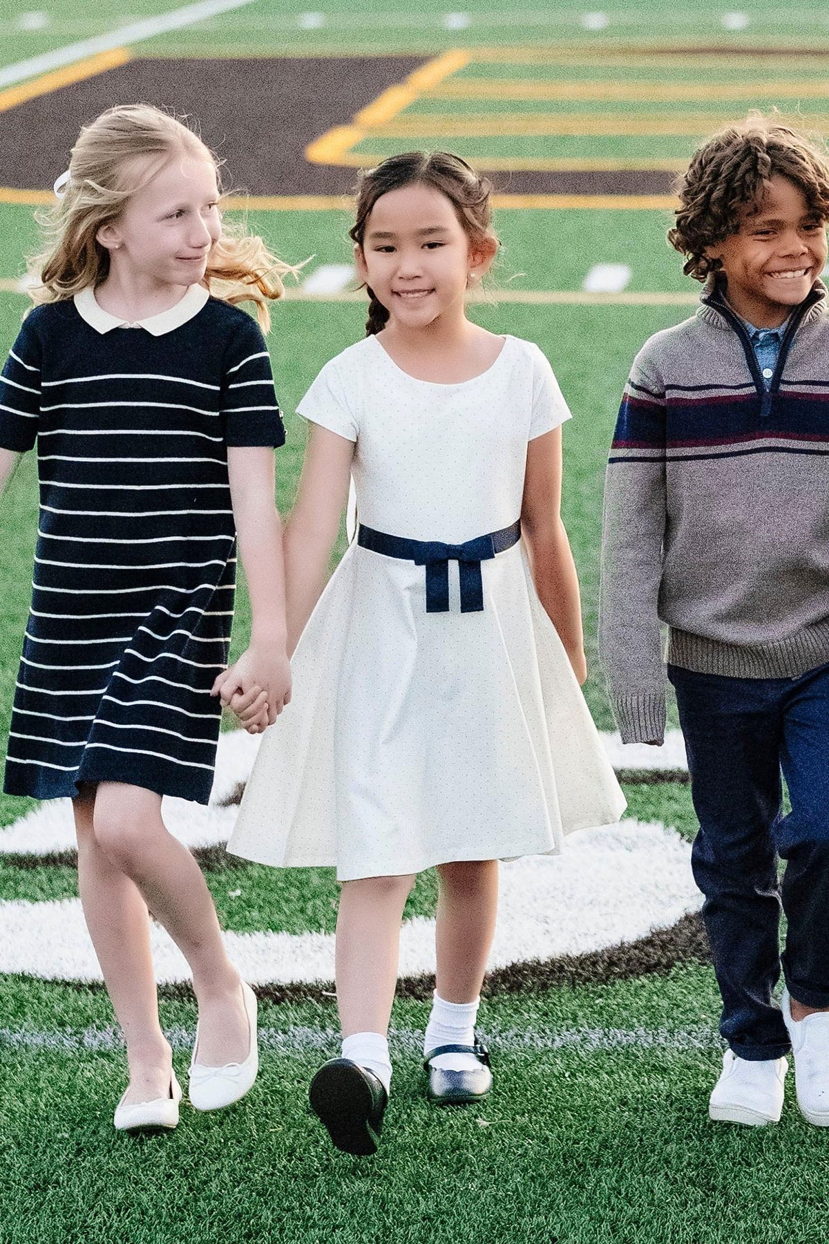 Three children walk hand in hand on a sports field; one wears a white dress with a navy bow.