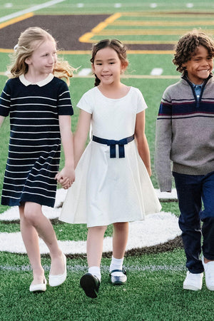 Three children walk hand in hand on a sports field; one wears a white dress with a navy bow.