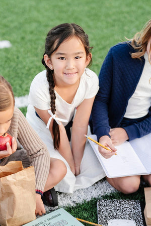 Young girl in a white pindot dress sits on the grass, smiling, with friends holding notebooks and snacks.
