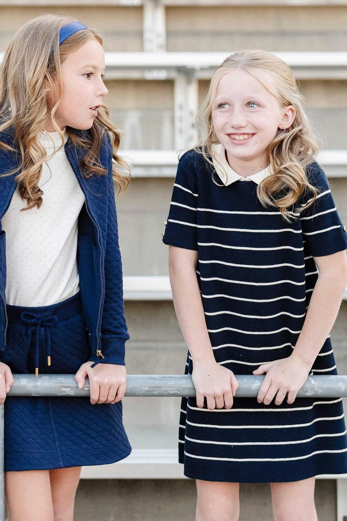 Two girls stand by a railing, one in a navy quilted jacket and the other in a striped dress, smiling.