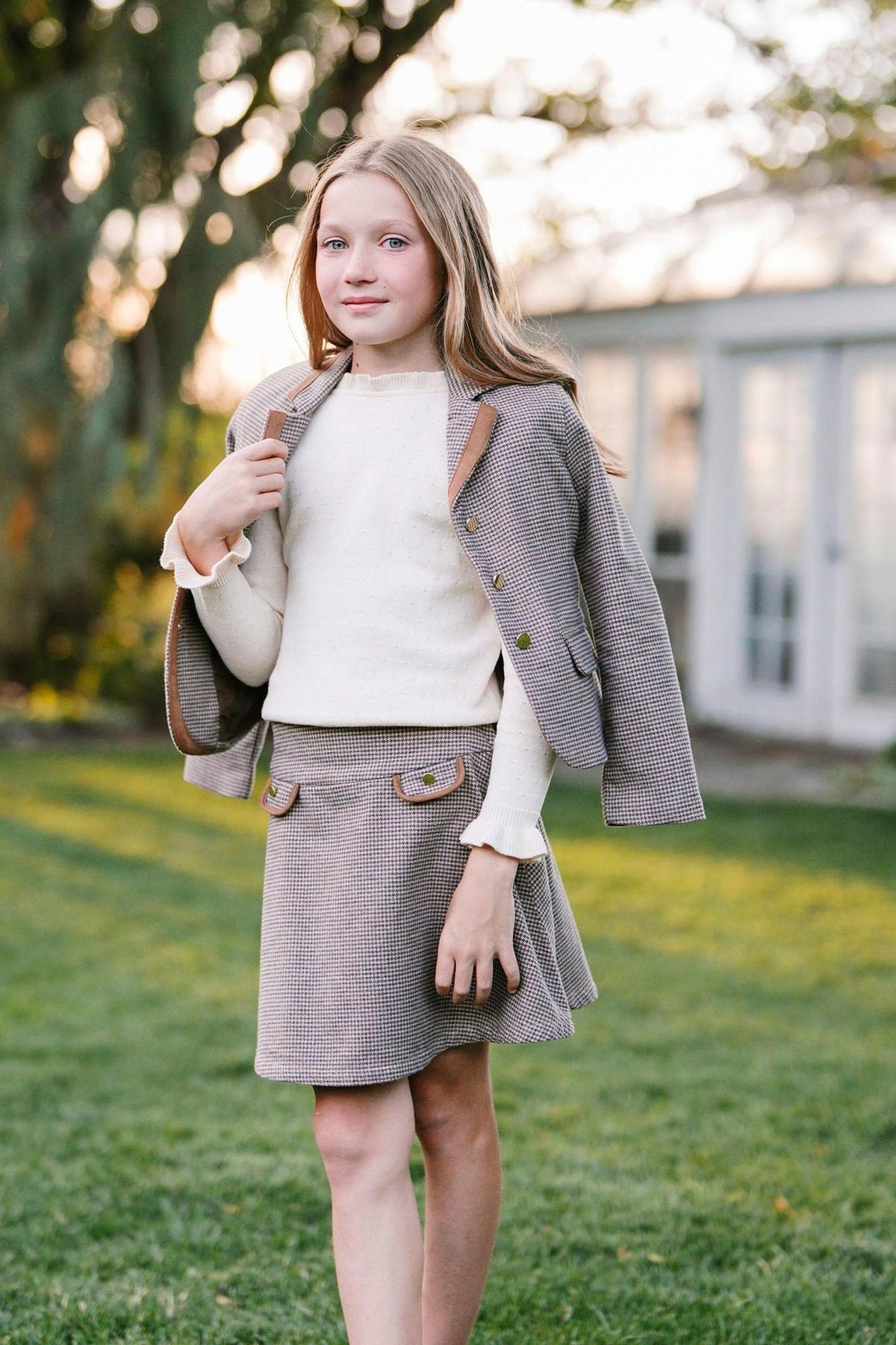 A young girl poses outdoors wearing a stylish houndstooth blazer and matching skirt, complemented by a cream sweater.