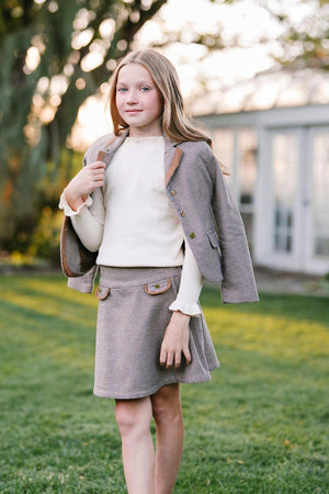 A young girl poses outdoors wearing a stylish houndstooth blazer and matching skirt, complemented by a cream sweater.