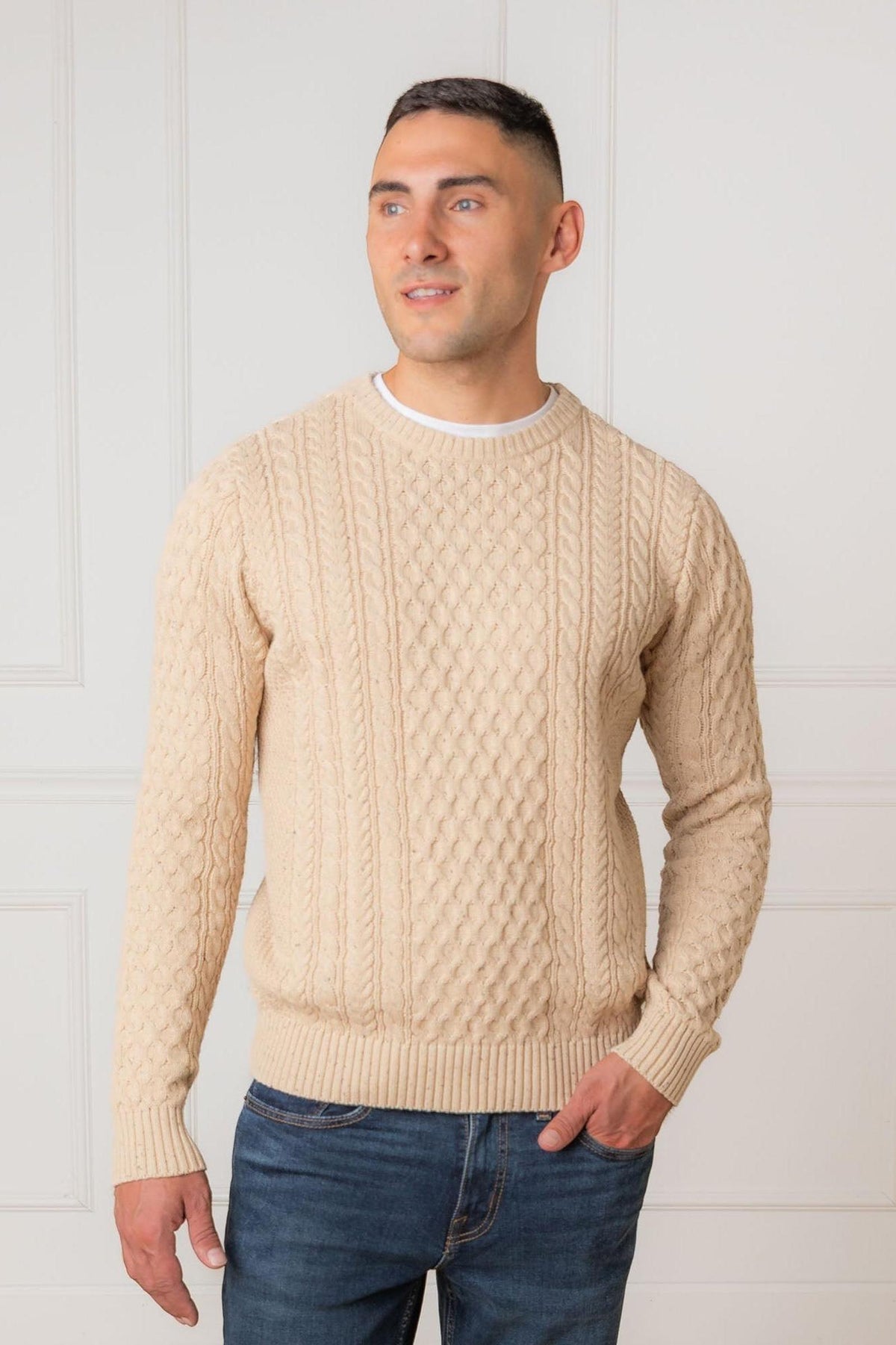 A man stands confidently in a beige organic cotton fisherman sweater featuring a textured cable knit design. The cozy crewneck pullover is set against a simple, light-colored background.