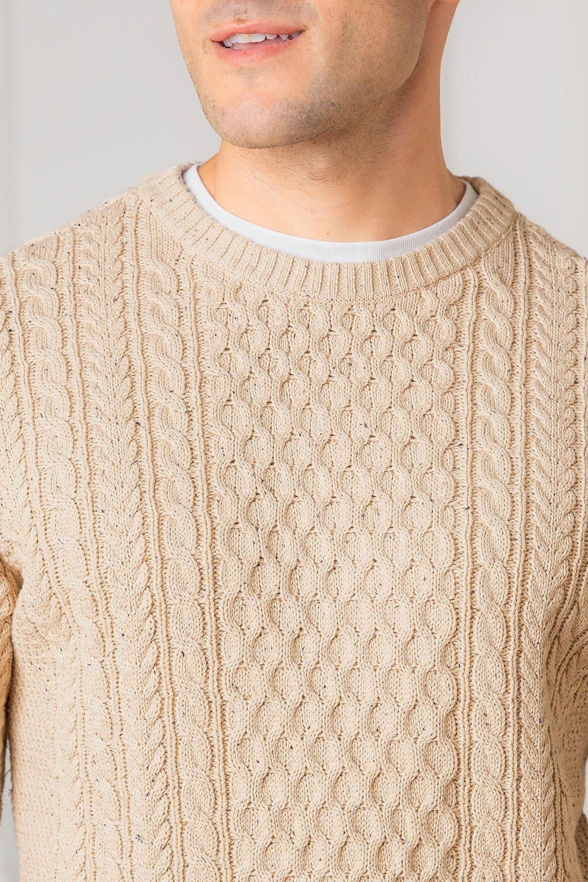 A close-up of a man wearing a natural fleck organic cotton fisherman sweater, featuring a textured allover cable design and classic crewneck style. The sweater showcases a warm, neutral color.