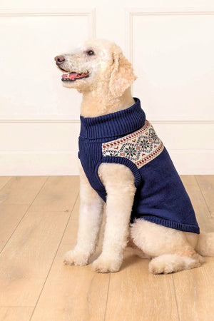 A light-colored dog wears a cozy navy Fair Isle sweater with a ribbed collar, sitting on a wooden floor.