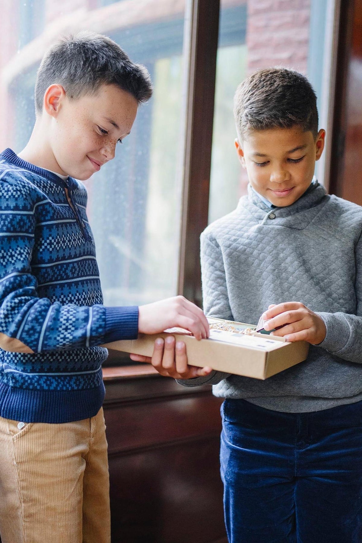 Two boys are exchanging a gift box indoors, one wearing a navy patterned sweater and the other in a gray.
