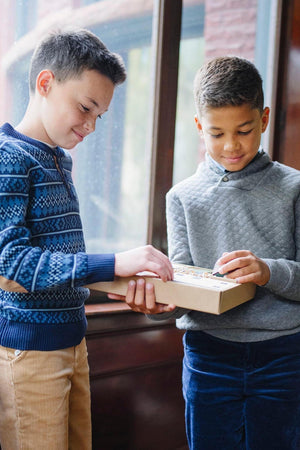 Two boys are exchanging a gift box indoors, one wearing a navy patterned sweater and the other in a gray.