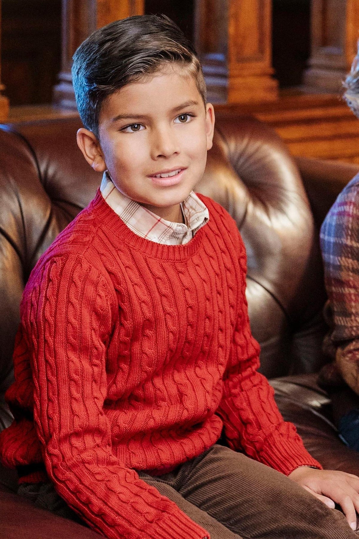 A boy wearing a red cable knit sweater sits on a brown leather couch, smiling and looking off-camera.