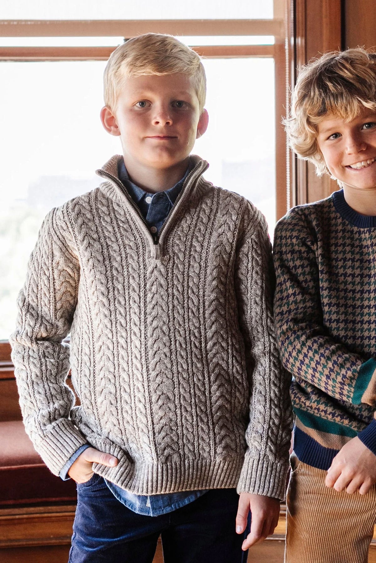 A boy wears a taupe cable knit sweater with a half zip, standing beside another boy in a patterned sweater.