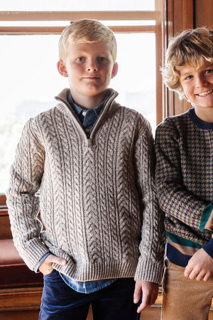 A boy wears a taupe cable knit sweater with a half zip, standing beside another boy in a patterned sweater.