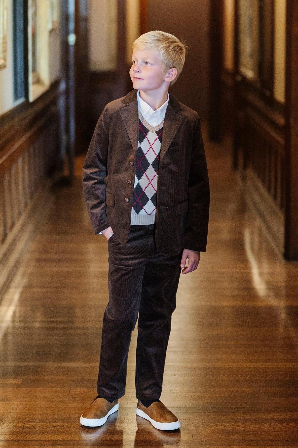 A young boy stands in a hallway wearing chocolate corduroy pants, a patterned sweater vest, and slip-on shoes.