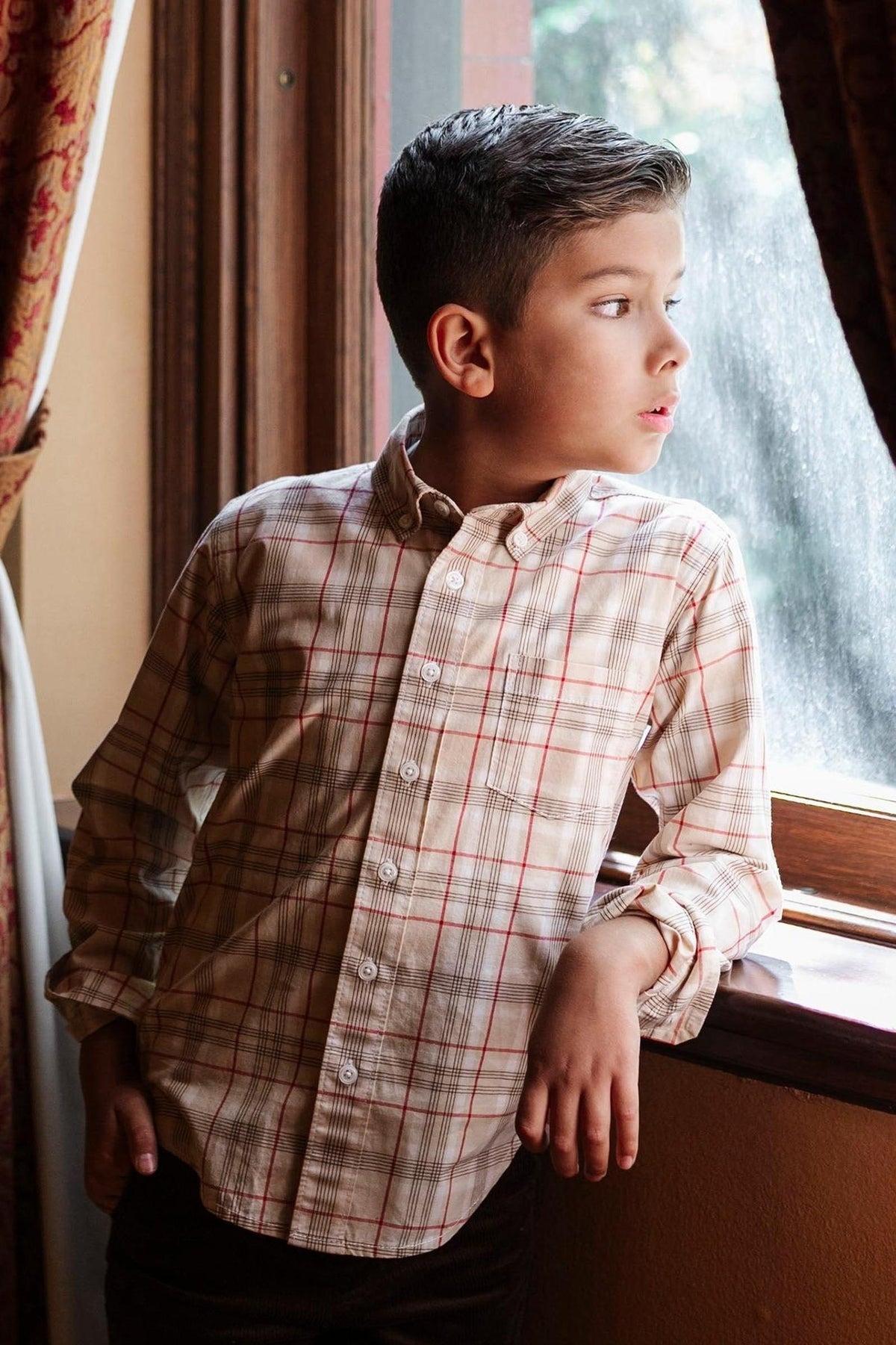 A young boy wearing a plaid button-down shirt leans against a windowsill, gazing thoughtfully outside.