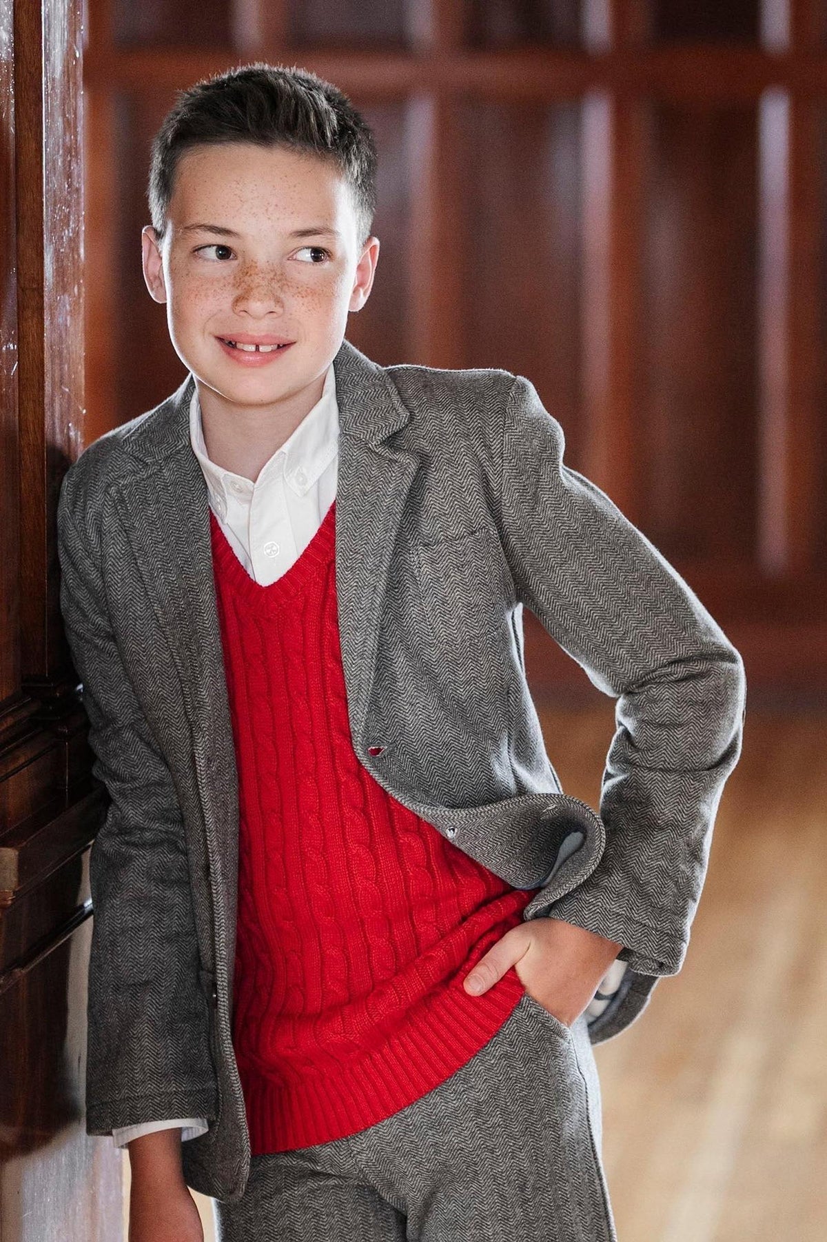 A young boy smiles while leaning against a wooden wall, wearing a red cable sweater vest over a white shirt.