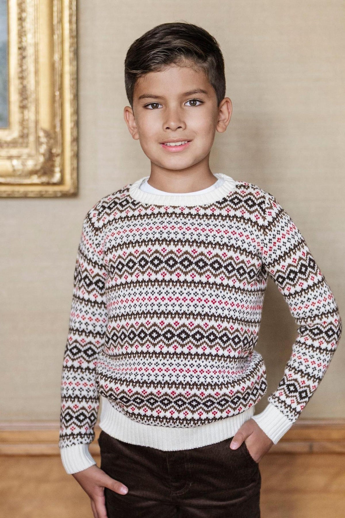 A young boy stands smiling in a cozy, patterned sweater featuring a Fair Isle design in red, black, and cream. The sweater has a crew neck and ribbed cuffs.
