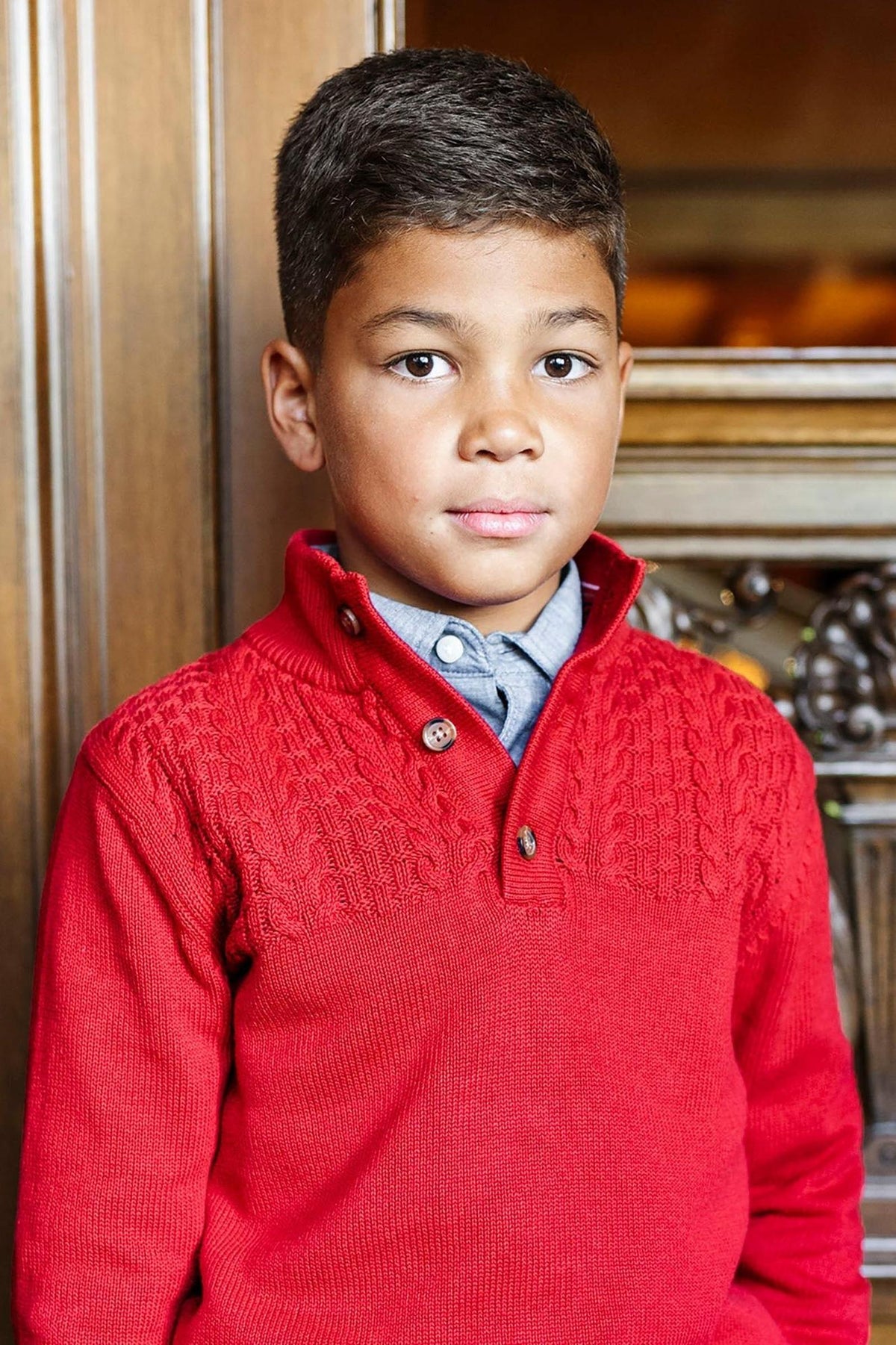 A young boy wears a vibrant red mock neck sweater with subtle textured knit details, standing against a wooden backdrop.