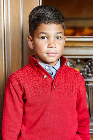 A young boy wears a vibrant red mock neck sweater with subtle textured knit details, standing against a wooden backdrop.