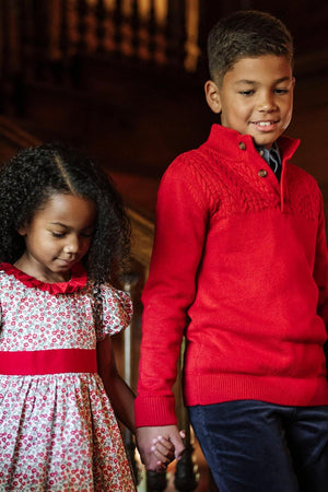 A boy in a red mock neck sweater holds hands with a girl wearing a floral dress with a red.