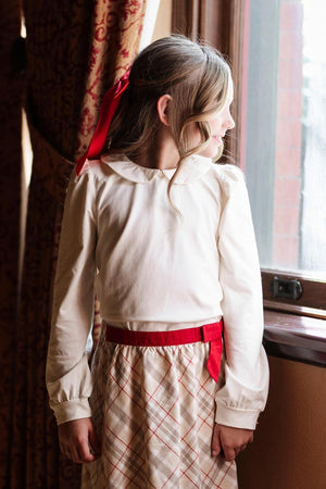 A girl wearing an ivory Peter Pan collar knit top and a plaid skirt with a red belt stands by.