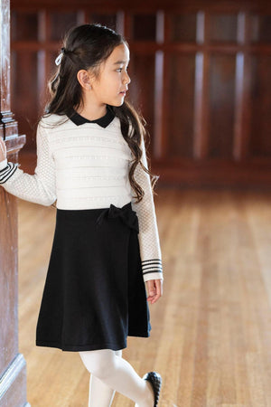 A young girl in a stylish white and black dress with a Peter Pan collar and a bow at the waist walks gracefully on polished wooden floors.
