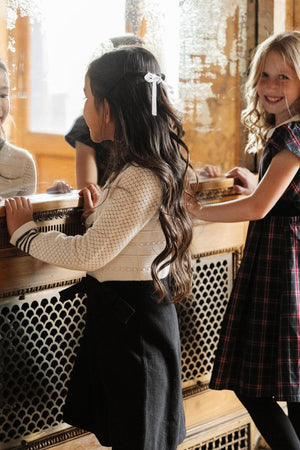 Two young girls stand in front of a vintage mirror, one wearing a cream sweater with textured stitches and a black skirt, while the other dons a plaid dress.