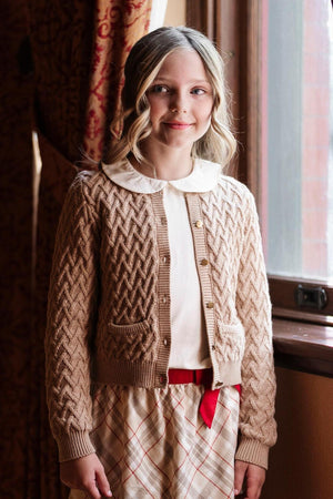 A young girl stands by a window wearing a camel-colored herringbone cardigan made of organic cotton, featuring a collared shirt and a plaid skirt, exuding a classic style.