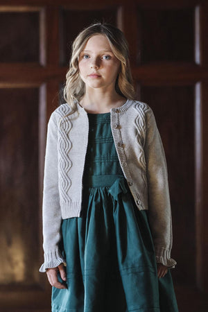 A young girl wears a light gray cable-knit cardigan over a dark green dress, posing against a wooden backdrop.