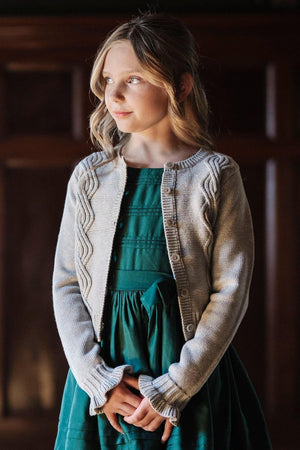 A girl with wavy hair wears a gray cable-knit cardigan over a green dress, showcasing ruffle cuffs and a button.