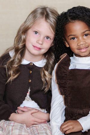 Two girls sit close together, one in a chocolate brown cropped cardigan and the other in a brown corduroy dress.