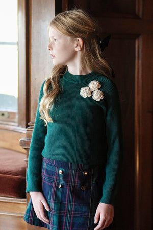 A girl stands by a window wearing a green ribbed sweater with three decorative rosettes, paired with a plaid skirt.