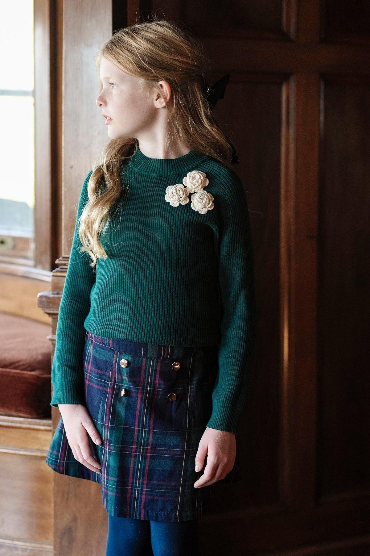 A young girl stands in profile, wearing a green ribbed sweater with decorative floral appliqués and a navy/green plaid skirt featuring double gold buttons.
