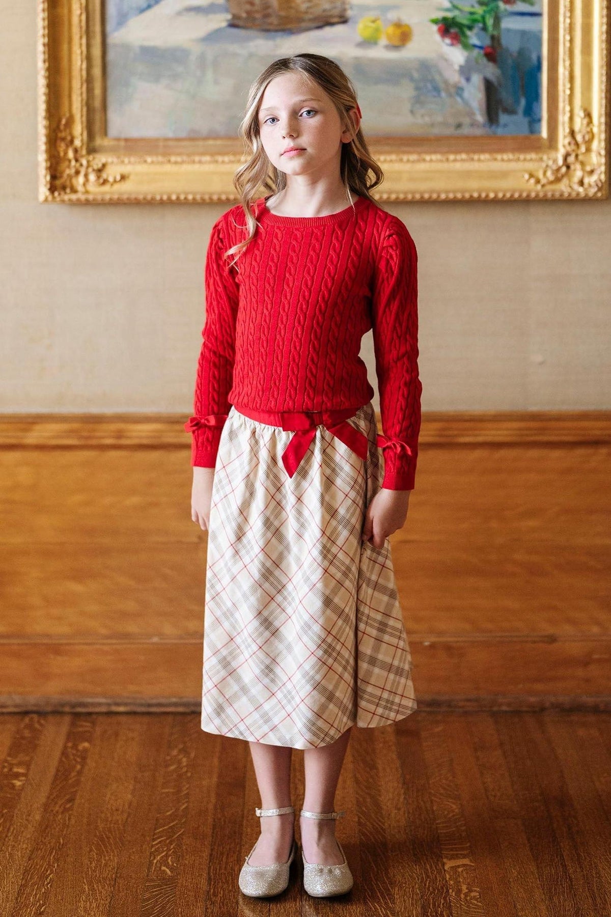 A girl in a red cable-knit sweater and plaid skirt stands against a wooden backdrop, wearing sparkly shoes.
