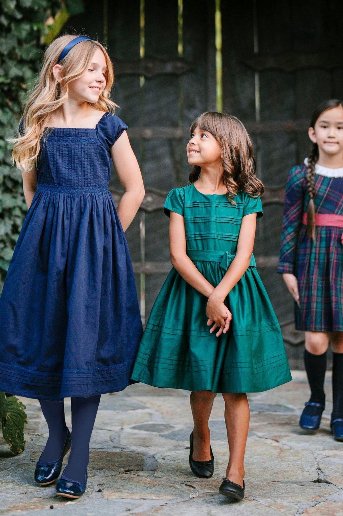 Three girls are smiling and wearing elegant dresses: one in navy, one in green, and another in plaid, set against.