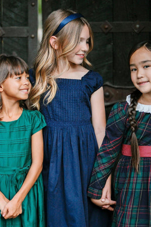 Three young girls are smiling, wearing elegant dresses: a navy puff sleeve dress, a green striped dress, and a plaid.