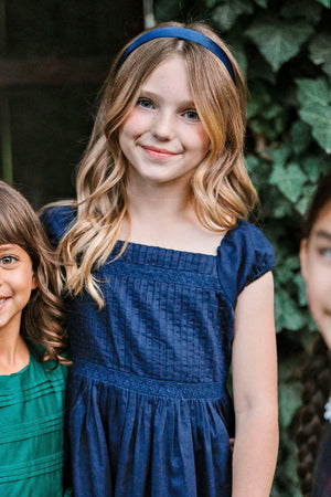 A girl with wavy blonde hair wears a navy puff sleeve dress, smiling against a backdrop of greenery.