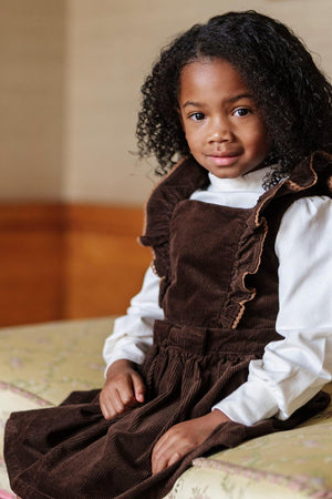 A young girl with curly hair wears a brown corduroy jumper dress with ruffled sleeves, sitting on a floral-patterned surface.