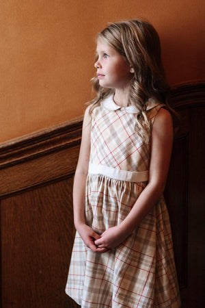 A young girl stands by a wooden wall, wearing a sleeveless ivory plaid dress with a Peter Pan collar.