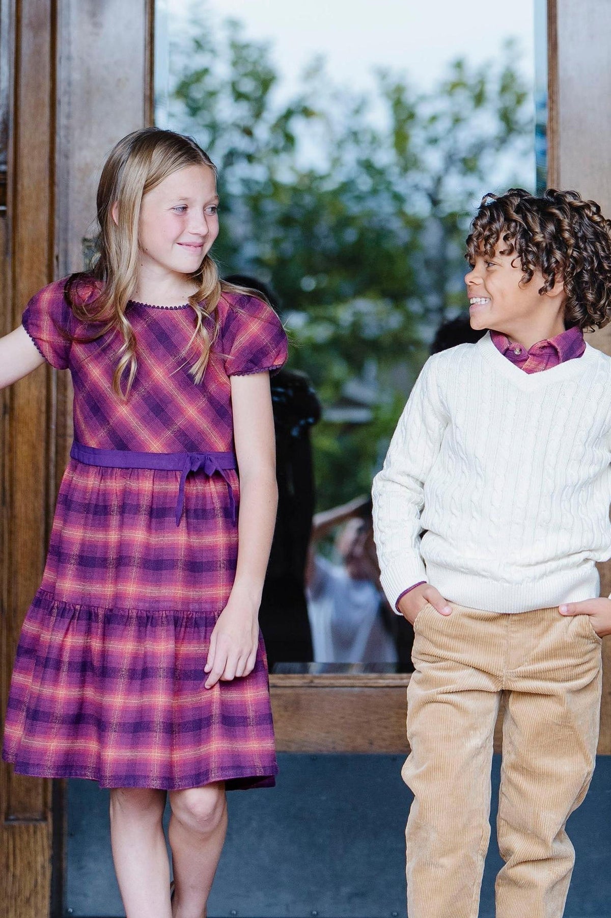 A girl in a sugar plum plaid dress smiles at a boy wearing a white cable-knit sweater and tan pants.