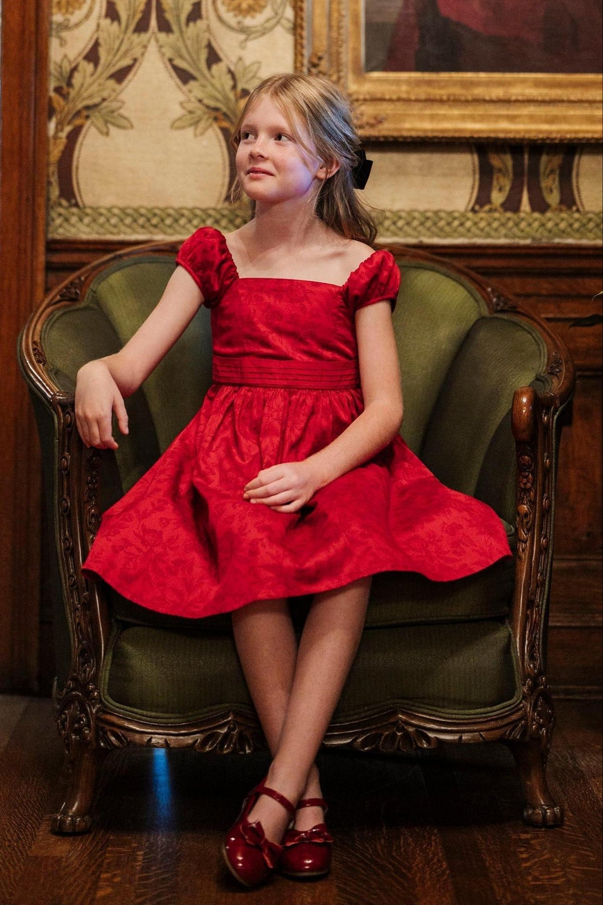 A young girl sits gracefully on a vintage green armchair, wearing a vibrant red dress with puff sleeves and a fitted bodice, exuding a classic and elegant charm.