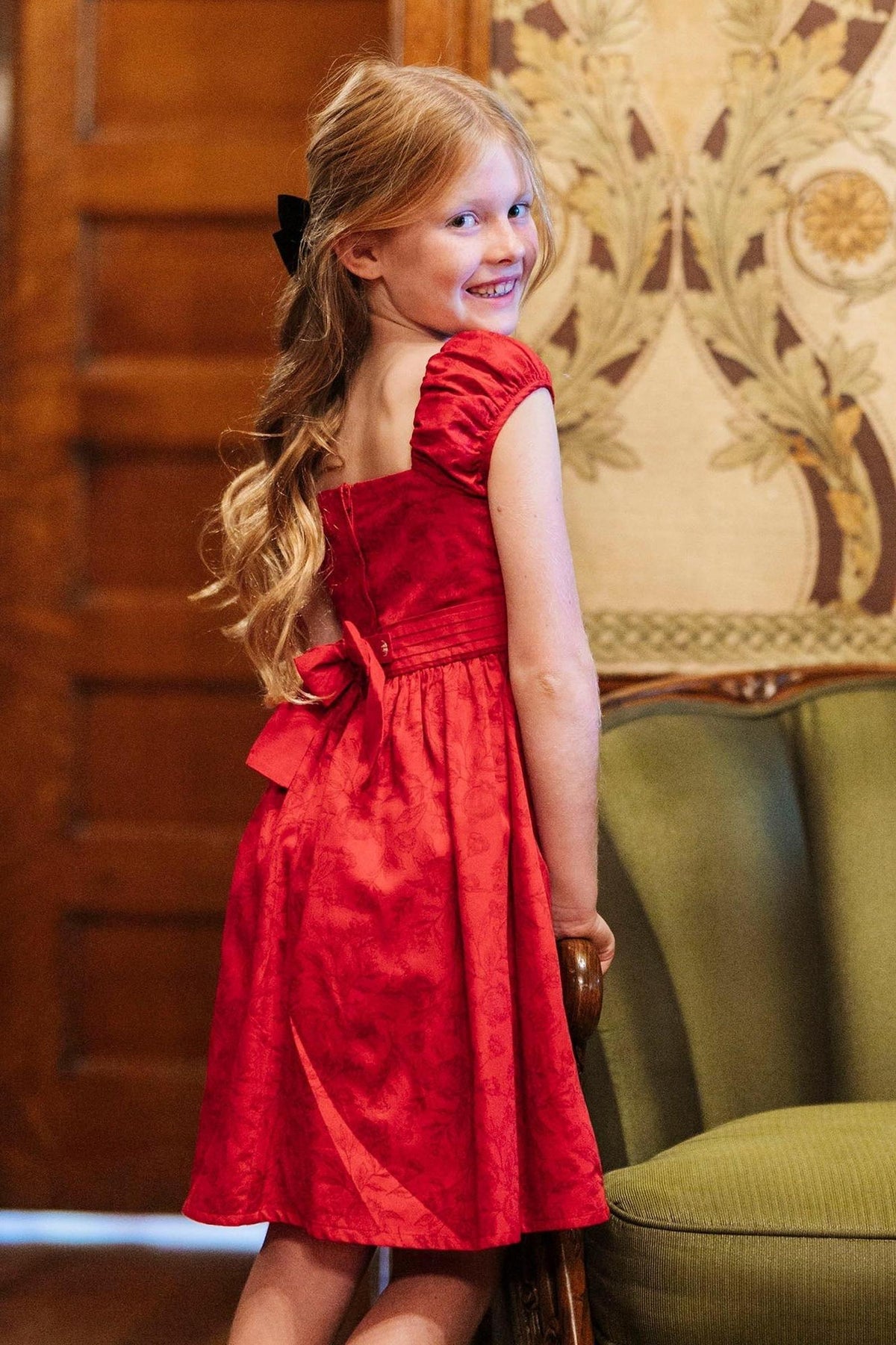 A young girl smiles while wearing a fireside red toile dress with puff sleeves and a bow at the waist.