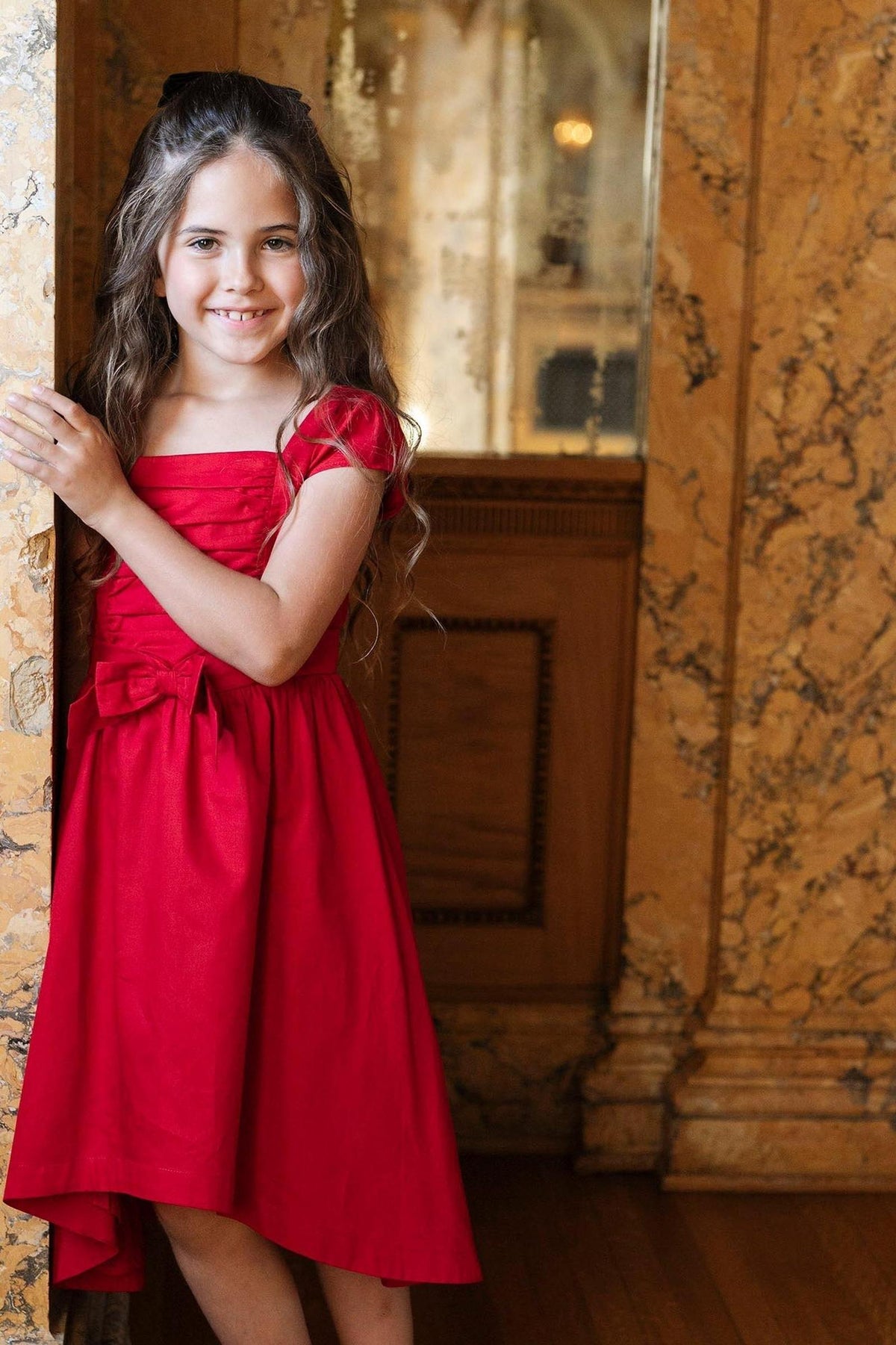 A girl in a red high-low dress with cap sleeves and a bow stands near a marble wall, smiling brightly.